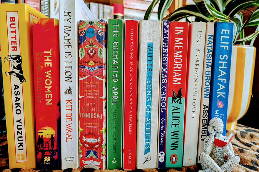 Books arranged on a shelf with colorful spines, a small reading statue, and a potted plant.